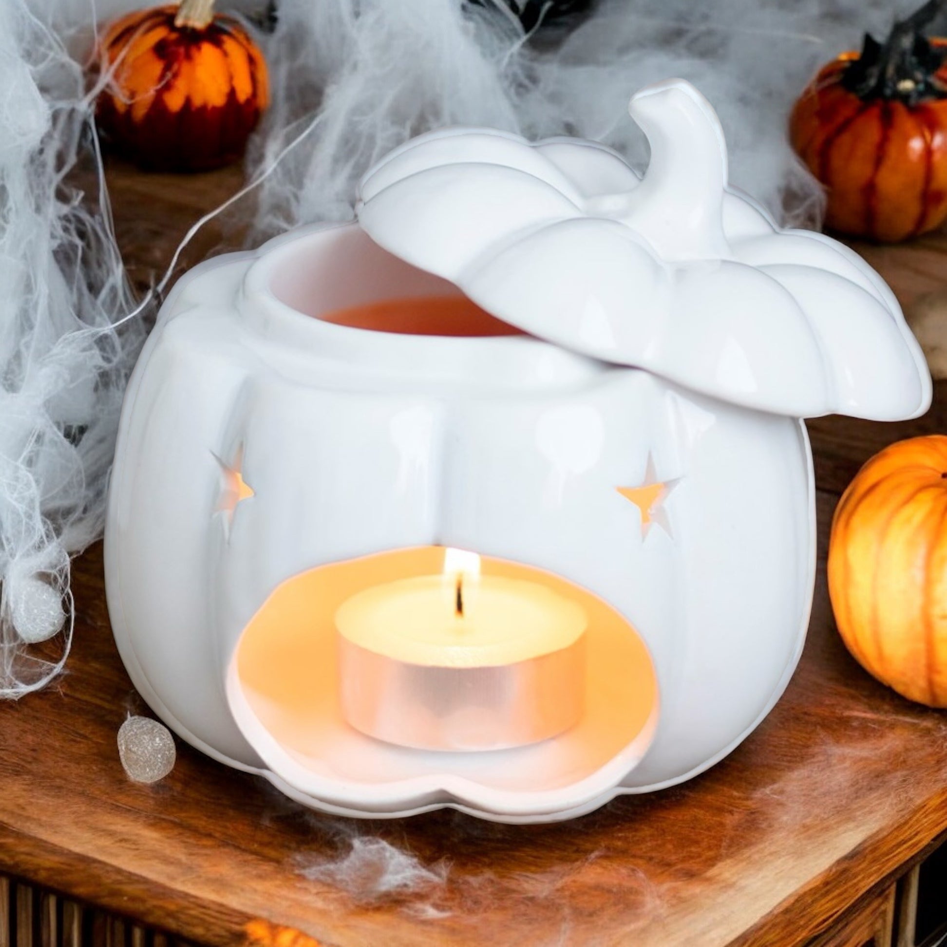 A white pumpkin-shaped oil burner with a tealight candle inside, placed on a wooden surface with decorative spider webs and small orange pumpkins around.